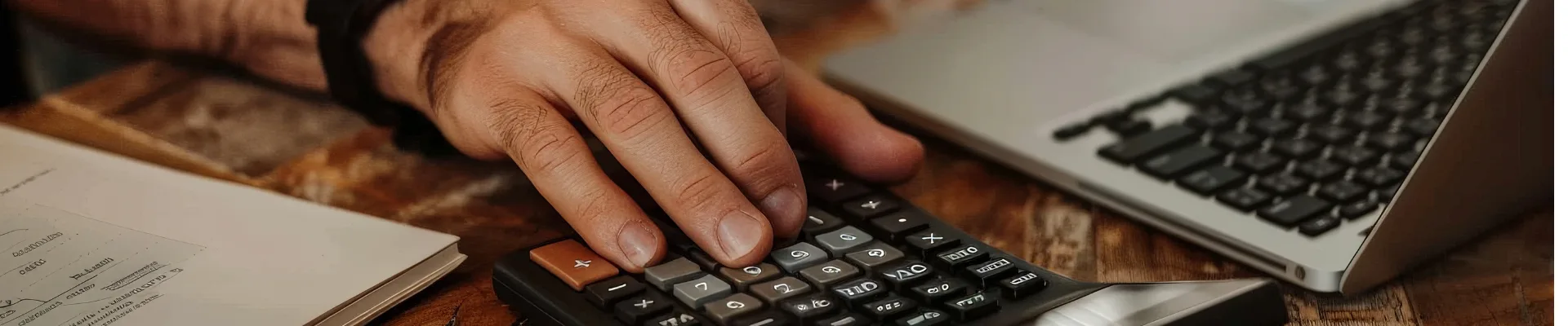 Person using a calculator at a wooden desk with a laptop, smartphone, and financial documents.