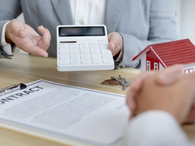 Professional advisor holding a calculator, house model, keys, and contract document visible on the table.